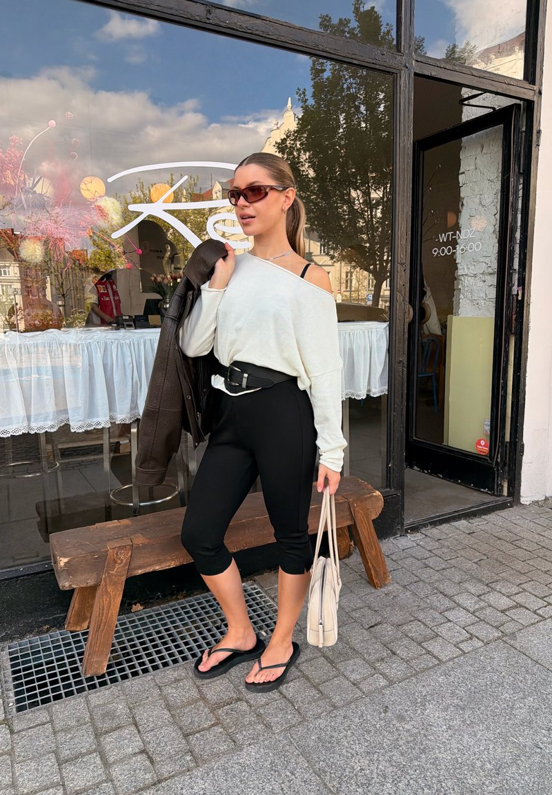 Jeune femme en lunettes de soleil tenant une veste en cuir sur l'épaule et un sac cabas, debout dans une rue pavée devant un café avec une grande fenêtre.