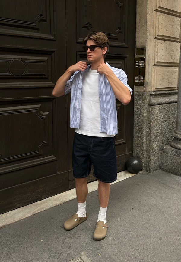 Casual outfit met een wit t-shirt, een gestreepte short-sleeve shirt, donkere denimshorts en beige klompen met witte sokken.