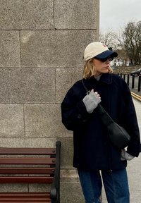 Femme en manteau sombre, jean bleu, casquette beige et marine, et lunettes de soleil, debout près d'un mur en pierre à côté d'un banc en bois sur un trottoir de la ville.