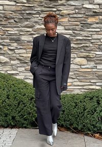 Zwarte, op maat gemaakte blazer over een aansluitend zwart topje, cargo-stijl zwarte broek en zilveren enkelboots. Achtergrond van een gestructureerde stenen muur.