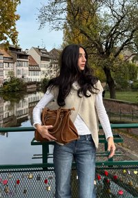 Strikket beige vest over en hvit langermet skjorte, blå jeans, holder en brun semsket veske. Grønn bro med hengelåser i bakgrunnen.