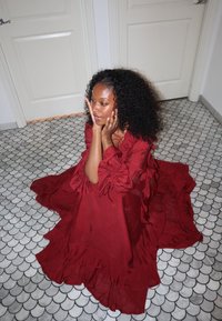 Femme aux cheveux bouclés portant une robe rouge fluide, assise sur un sol en carreaux à motifs, reposant son visage sur ses deux mains près de portes blanches fermées.