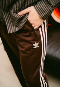 Calças de treino castanhas da adidas com riscas brancas nas laterais, apresentando um bolso com fecho e um detalhe de logótipo. Usadas com uma camisa preta às riscas.