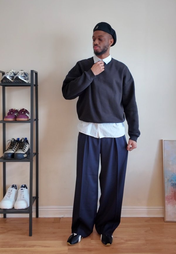 Homme ajustant son col portant un béret noir, un pull noir sur une chemise blanche, un pantalon large bleu marine, debout près d'un porte-chaussures avec cinq paires de chaussures.