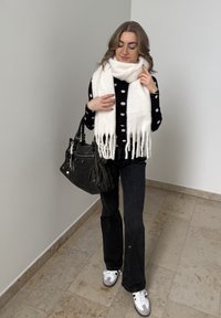 Femme debout à l'intérieur portant un pull noir à pois, une écharpe blanche, un jean noir, des baskets blanches et portant un grand sac à main noir.