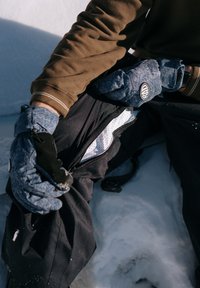 Moufles en tissu bleu avec poignets élastiques, pantalon de neige noir avec une poche zippée, portés sur des couches blanches texturées, posés sur la neige.