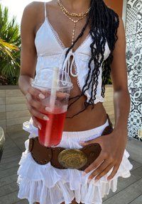 Valkoinen lyhyt toppi pitsireunuksella, kerroksellinen valkoinen volangihame, ruskea vyö koristeellisella kultaisella soljella, pitämässä kirkasta juomaa, jossa on punaista nestettä.