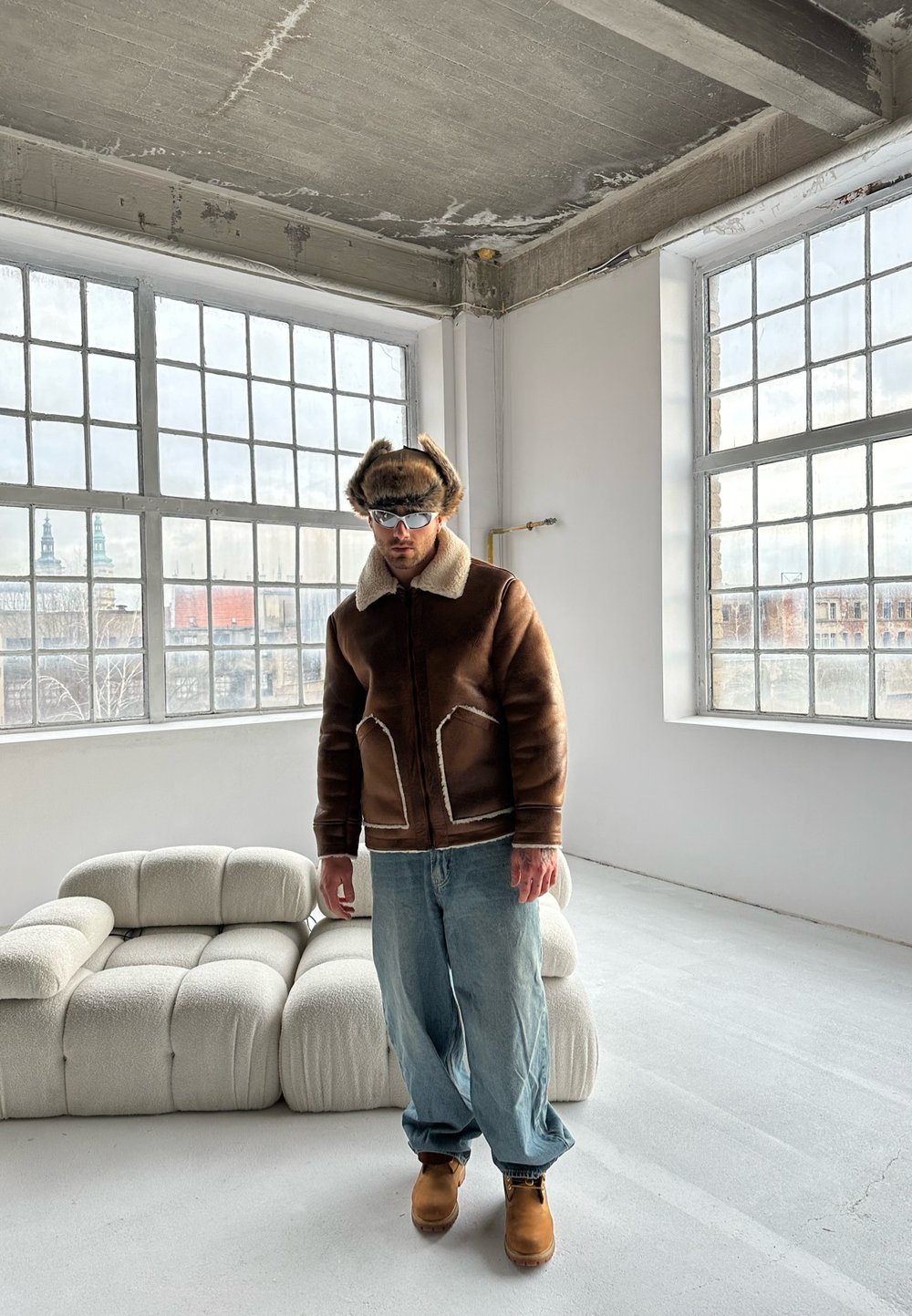 Giacca in shearling marrone con cuciture bianche, cappello di pelliccia marrone, jeans blu e stivali beige. Interni moderni con grandi finestre e mobili bianchi.