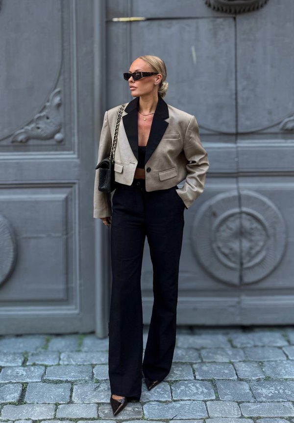 Donna con capelli biondi raccolti in uno chignon basso, che indossa occhiali da sole neri, un blazer beige corto, una maglietta nera, pantaloni neri a vita alta e scarpe appuntite.