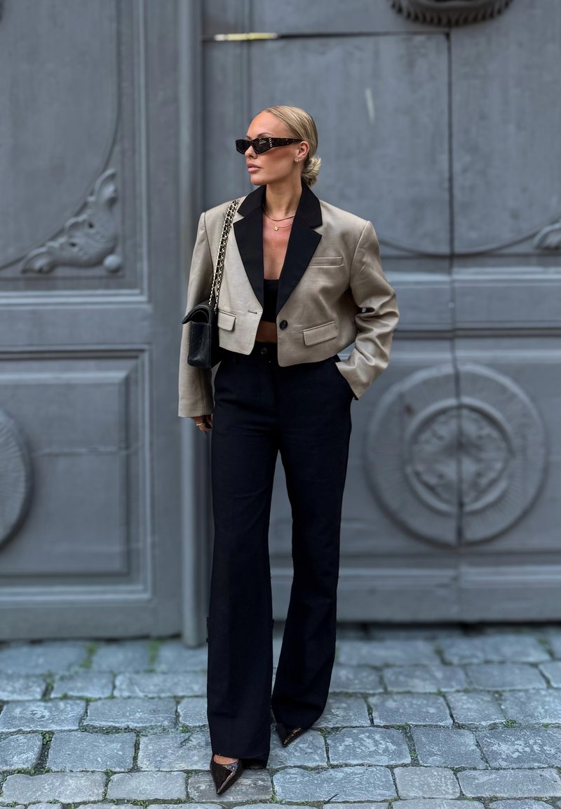 Femeie cu păr blond prins într-un coc jos, purtând ochelari de soare negri, un sacou scurt bej, un top negru, pantaloni negri cu talie înaltă și pantofi ascuțiți.