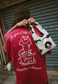 Camiseta de algodón roja con gráfico blanco de un personaje de dibujos animados y texto; bolsa de patrón en blanco y negro con asa y logo.