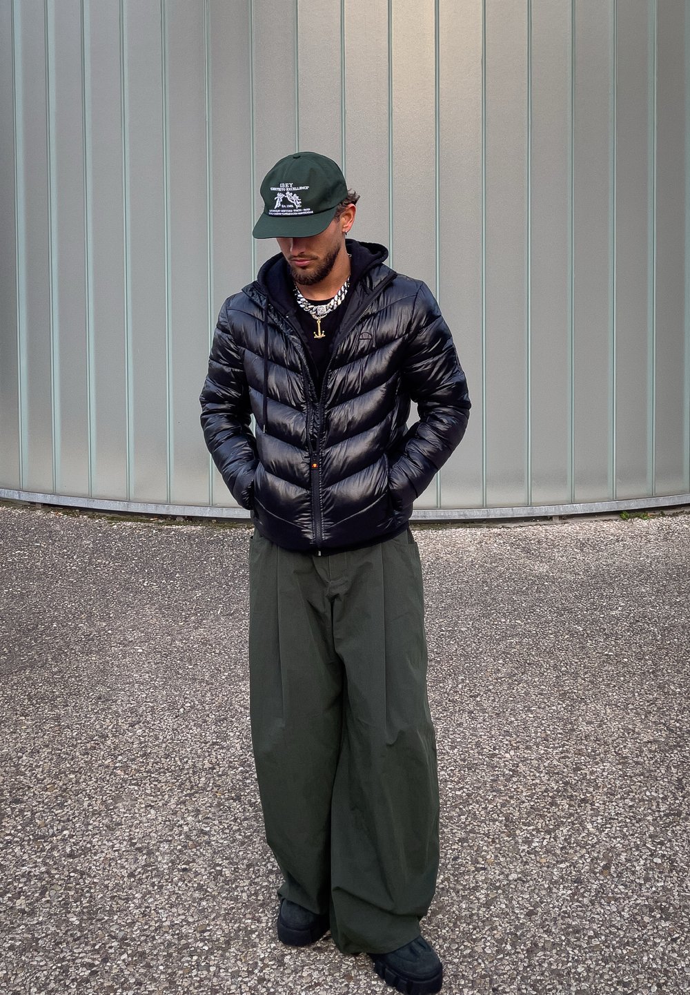 Homme portant une doudoune noire, un pantalon large vert foncé, des chaussures noires et une casquette verte, debout les mains dans les poches contre un mur métallique courbé.