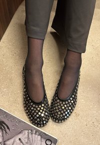 Άτομο που φοράει μαύρο διαφανές καλσόν και μαύρες μπαλαρίνες με ασημένιους κυκλικούς τρουκς, στέκεται σε μπεζ υφασμένο πάτωμα κοντά σε ένα περιοδικό.