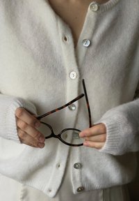 Biały, teksturowany kardigan z dużymi, perłowymi guzikami, ściągacze na rękawach, trzymany przez jasne dłonie z tortoisowymi okularami.