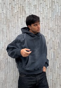 Jeune homme portant un hoodie noir ample et un jean noir, debout contre un mur de briques grises texturé, regardant sur le côté avec une main dans la poche.