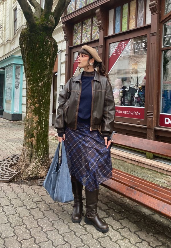 Moteris dėvi rudą odinę striukę, tamsiai mėlynos languotos sijoną, rudus batus, smėlio spalvos beretę, laiko mėlyną rankinę, stovi ant šaligatvio prie parduotuvės vitrinų ir medžio.