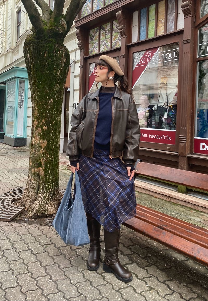 Femme portant une veste en cuir marron, une jupe marine à carreaux, des bottes marron, un béret beige, tenant un sac bleu, debout sur le trottoir près d'une vitrine et d'un arbre.
