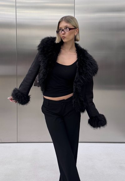 Chaqueta negra corta con cuello y puños de piel sintética, combinada con una blusa y pantalones negros ajustados. Textura suave con una silueta elegante.
