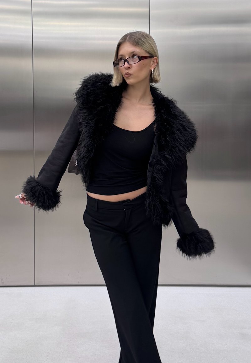 Schwarze kurz geschnittene Jacke mit Kunstfellkragen und -bündchen, kombiniert mit einem schwarzen eng anliegenden Oberteil und einer Hose. Glatte Textur mit einer eleganten Silhouette.