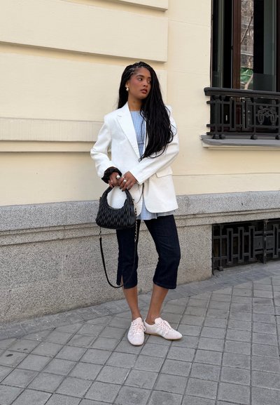 Mujer con cabello largo trenzado que lleva una blazer blanca, camisa de cuadros, pantalones cortos oscuros, zapatillas rosas, sosteniendo un bolso negro, de pie junto a una pared beige.
