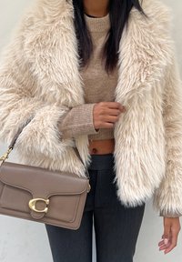 Chaquetón de piel sintética en crema con tejido texturizado, combinado con un suéter corto beige y pantalones gris de tiro alto; sosteniendo un bolso en taupe con accesorios dorados.
