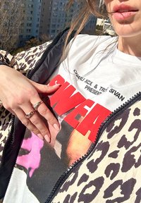Luipaardprint pufferjack met rits, boven een wit graphic t-shirt met dikke rode tekst. Hand versierd met ringen, met neutrale nagellak.