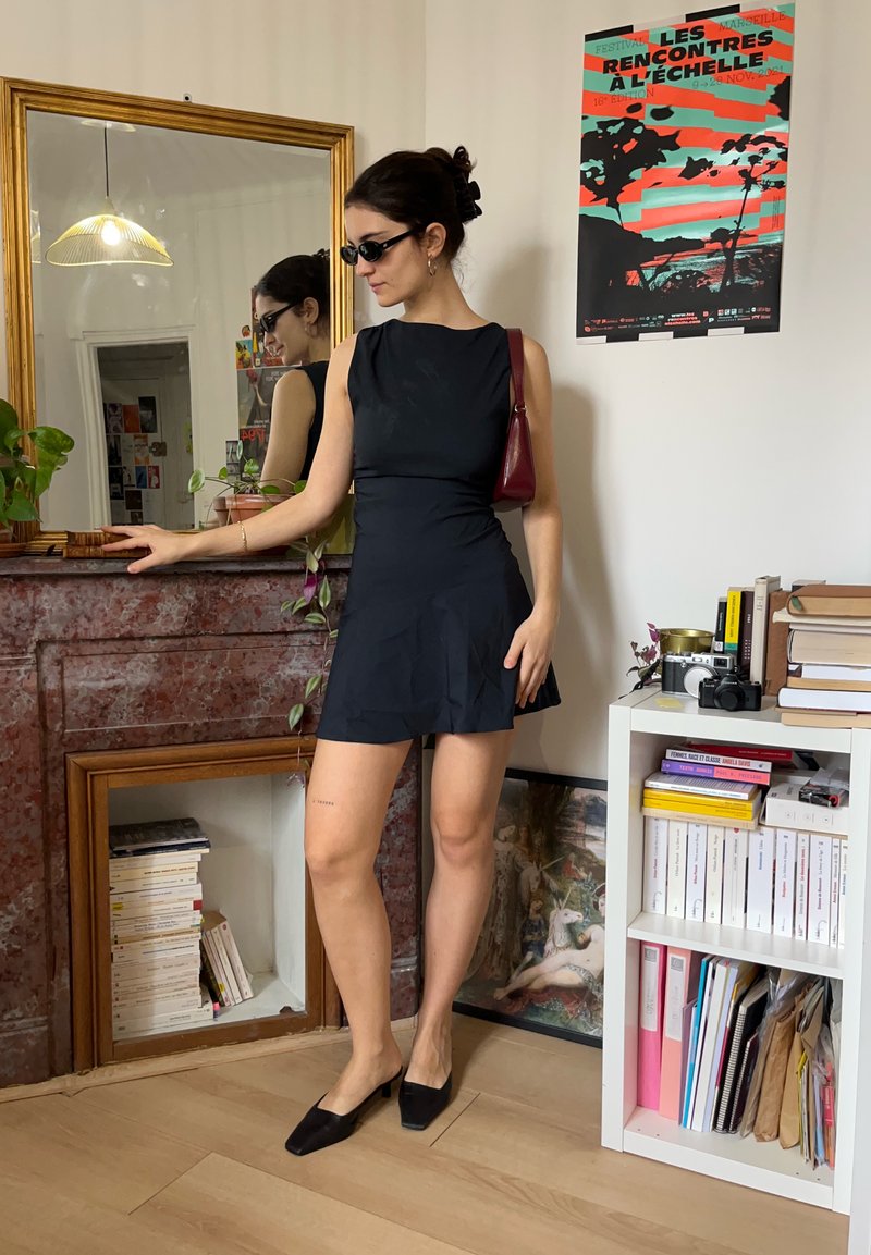 Femme en robe noire et lunettes de soleil se tient dans une pièce avec un miroir, une bibliothèque, un appareil photo vintage et une affiche colorée au mur.