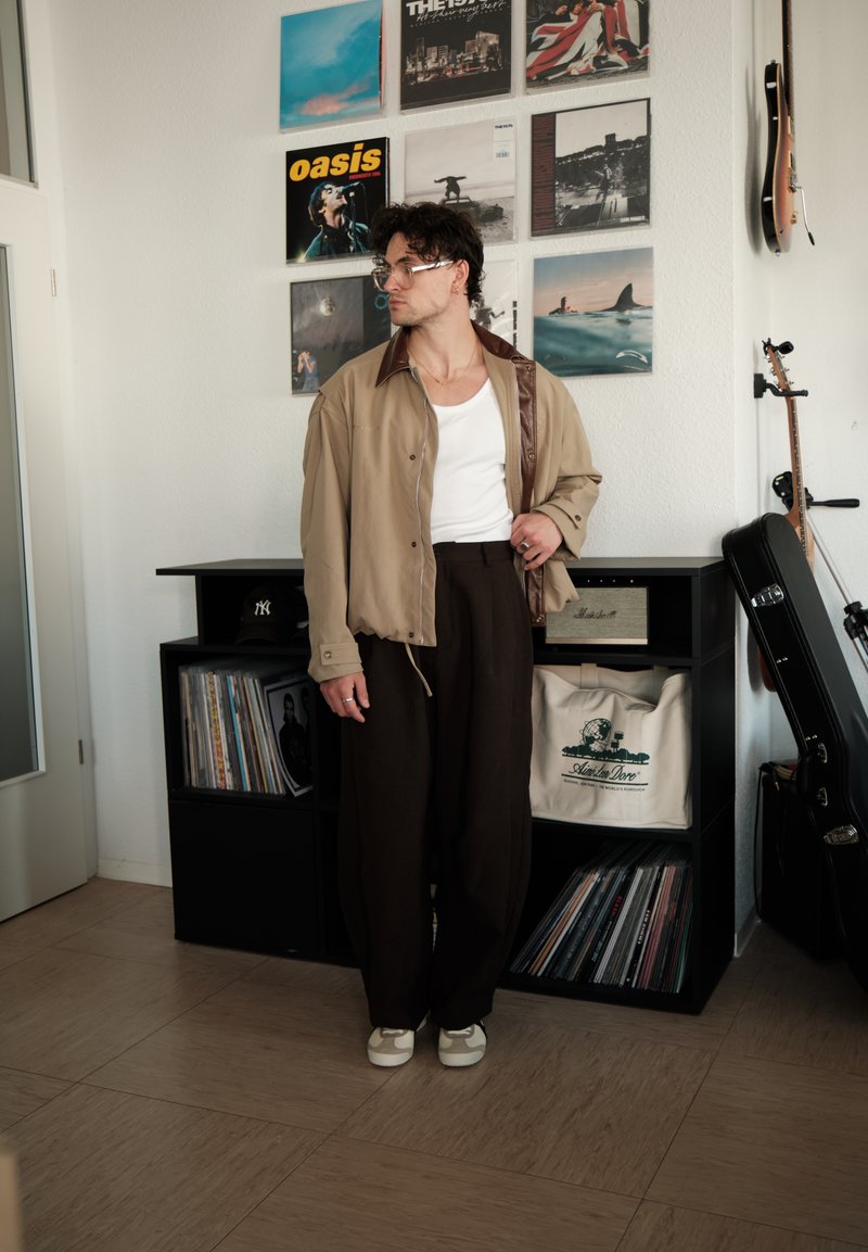 Homme portant des lunettes et une veste beige se tient près d'une platine vinyle, d'une collection de vinyles, de guitares et de pochettes d'album encadrées au mur.