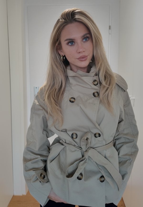 Une femme aux longs cheveux blonds pose en intérieur, vêtue d’un trench-coat croisé gris clair à ceinture et de créoles dorées.