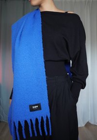 Blå teksturert skjerf drapert over en svart genser. Skjerfet har frynsete kanter og en etikett med teksten "ZIGN STUDIO." Ullaktig utseende, myk finish.