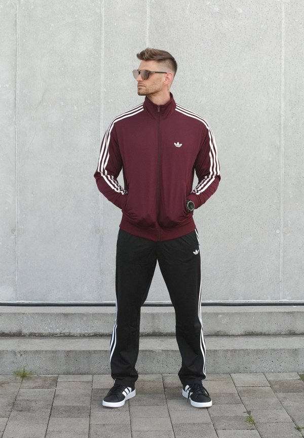 Muž ve vínově červené a černé sportovní soupravě Adidas s bílými pruhy, černých teniskách, slunečních brýlích a hodinkách na zápěstí stojí u šedé zdi.