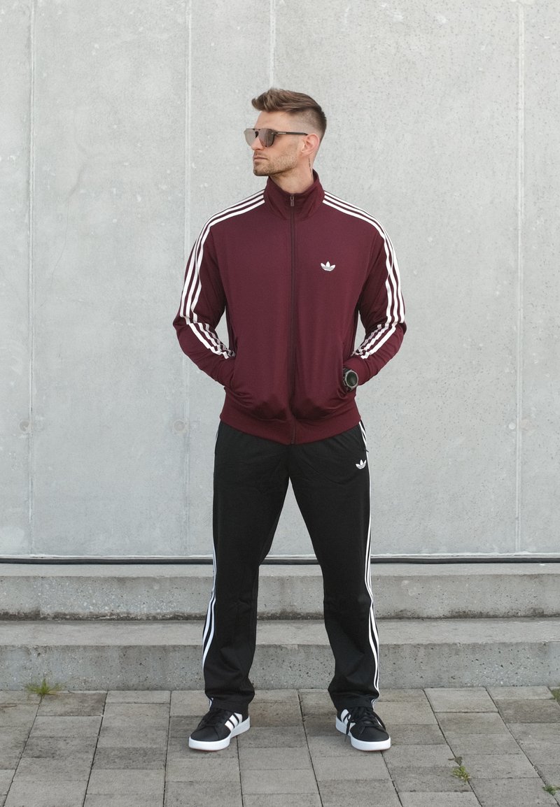 Homme portant un survêtement Adidas bordeaux et noir avec des bandes blanches, des baskets noires, des lunettes de soleil et une montre-bracelet, debout contre un mur gris.