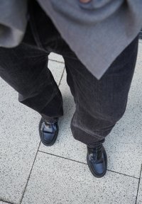 Persoon in zwarte leren loafers, donkere jeans en een grijze top die op lichtgrijze betegelde stoep staat.