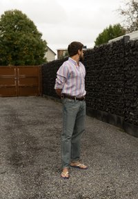 Cămașă din bumbac cu dungi în nuanțe pastel, blugi din denim bleu deschis și flip-flops cu curele albe și talpă albastră, pe un drum cu pietriș.