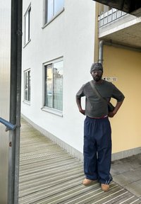 Homme noir portant une chemise à manches courtes grise, un pantalon bleu marine et des bottes marron clair, se tenant sur une passerelle en bois à côté d'un bâtiment blanc.