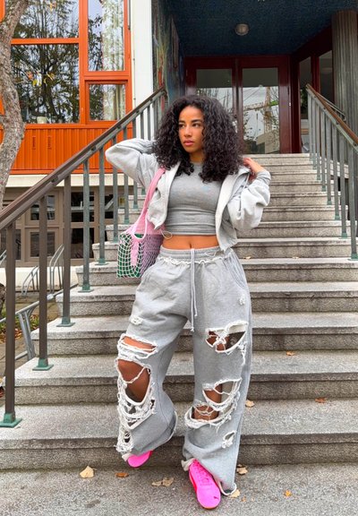 Pantalones de chándal grises desgastados con grandes cortes, combinados con un hoodie corto gris claro y zapatillas brillantes rosas. Accesorio: una bolsa de malla rosa.