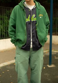 Grønn zip-up hettejakke over en mørk skjorte med knapper, kombinert med lysegrønne bukser. Funksjoner inkluderer hette, lommer og sporty detaljer.