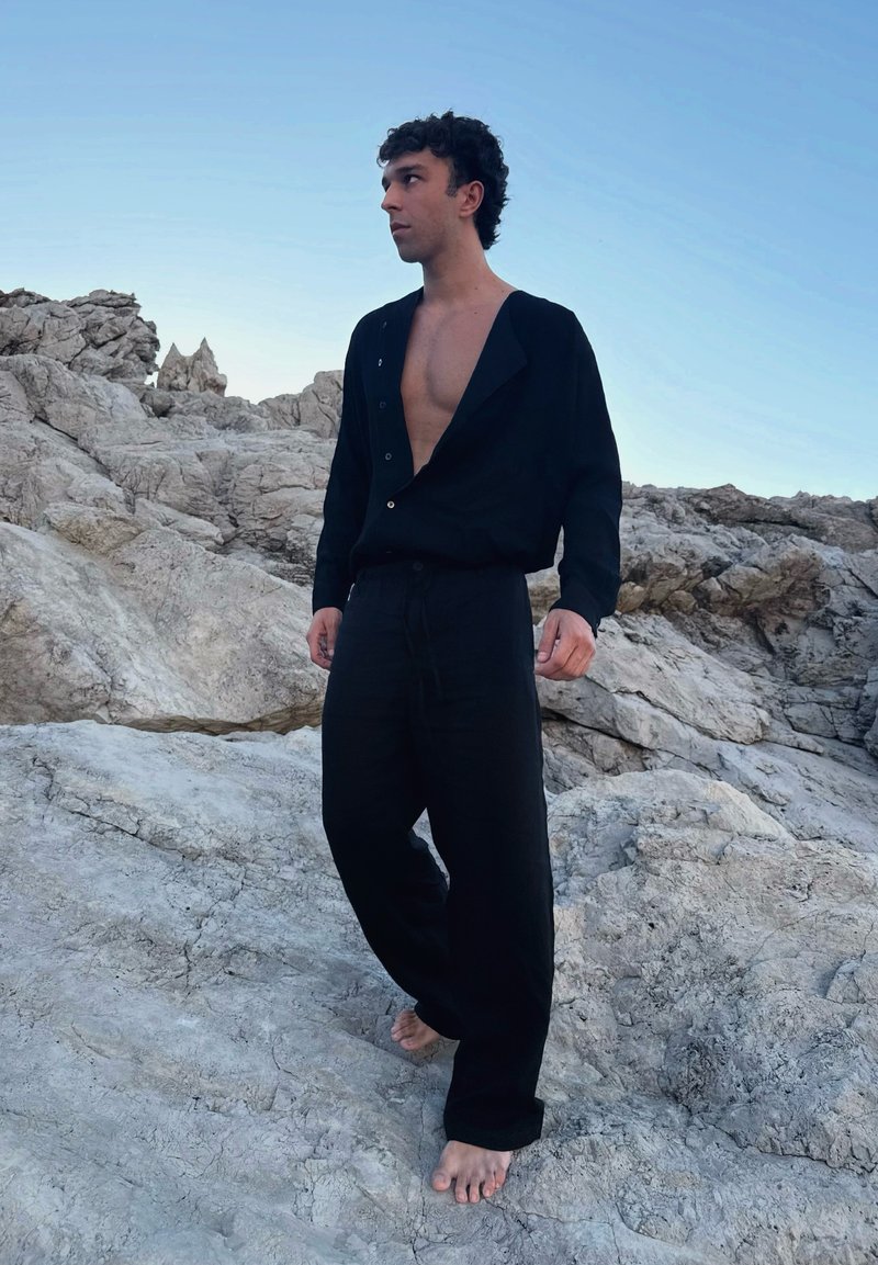 Homme aux cheveux bouclés portant un pantalon noir ample et une chemise noire ouverte, debout pieds nus sur un terrain rocheux sous un ciel clair.
