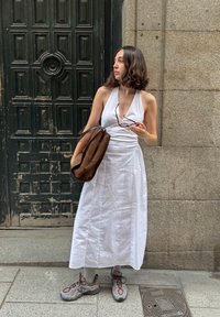 Biela halter šaty s plynulým dizajnom. Hnedá semišová kabelka cez rameno. Šedé športové topánky s ružovými akcentmi. Stojí vedľa textúrovaných tmavých dverí.