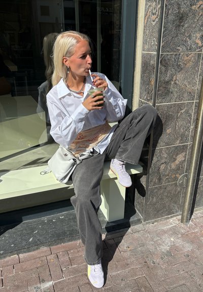 Una figura femenina está sentada en un borde, vistiendo una camisa blanca de gran tamaño, pantalones grises y zapatillas color lila, mientras bebe una bebida verde. Hay una bolsa blanca presente.