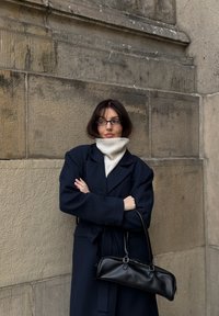 Manteau en laine bleu marine avec un col châle, superposé sur un col roulé blanc côtelé. Tenant un sac à main en cuir noir avec deux anses.