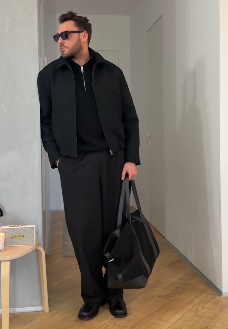 Homme en veste noire, pantalon et lunettes de soleil tenant un grand sac fourre-tout noir, debout à l'intérieur près d'une petite table avec un haut-parleur et une bougie.