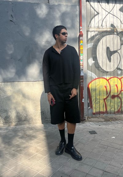 Camiseta negra de manga larga, pantalones cortos negros, calcetines negros y zapatos. De pie contra una pared gris con graffiti, sosteniendo un teléfono rojo.
