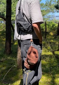 Borsa a tracolla in tessuto nero indossata a tracolla, con un cappello marrone attaccato in vita da una clip. Camicia a quadri grigia e jeans scuri.