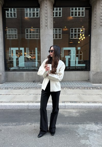 Chaqueta oversized de color beige claro, pantalones acampanados negros y botas negras de punta. Sosteniendo una taza de café, de pie frente a un edificio de cristal.