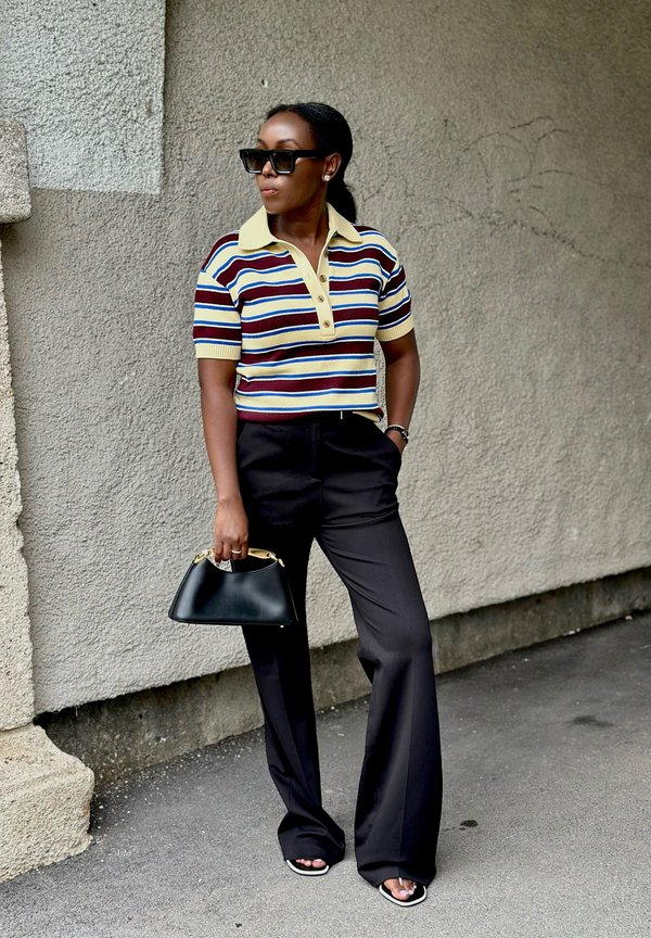 Donna con occhiali da sole neri, polo a righe a maniche corte, pantaloni neri a gamba larga, che tiene una borsa nera, in piedi contro un muro con texture.