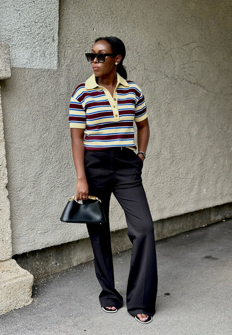 Woman wearing black sunglasses, striped short-sleeve polo shirt, black wide-leg pants, holding black handbag, standing against textured wall.