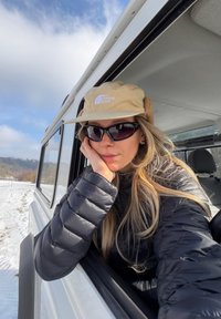 Νεαρή γυναίκα με μαύρο μπουφάν, μπεζ καπέλο και γυαλιά ηλίου, που ακουμπάει έξω από το παράθυρο ενός λευκού οχήματος, με χιονισμένο τοπίο και συννεφιασμένο ουρανό στο φόντο.