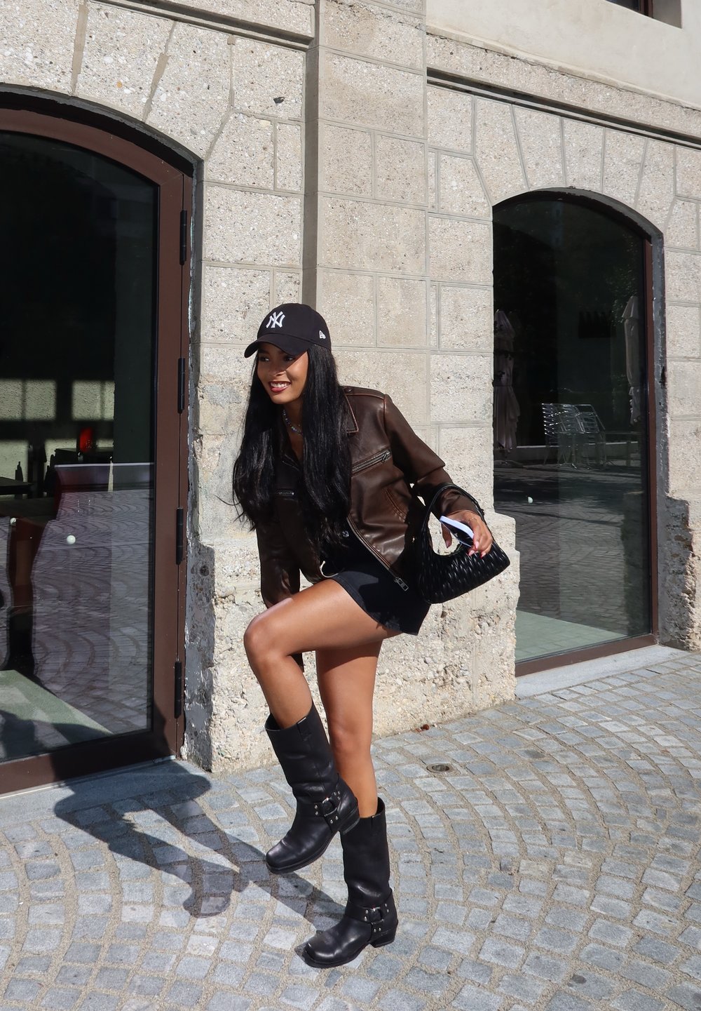 Καφέ δερμάτινο μπουφάν, μαύρο καπέλο με λογότυπο, μαύρη μίνι φούστα, μαύρες μπότες μέχρι το γόνατο με αγκράφες και μι�α πλεχτή μαύρη τσάντα. Φόντο από πέτρινο τοίχο.