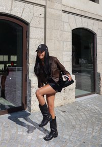 Καφέ δερμάτινο μπουφάν, μαύρο καπέλο με λογότυπο, μαύρη μίνι φούστα, μαύρες μπότες μέχρι το γόνατο με αγκράφες και μια πλεχτή μαύρη τσάντα. Φόντο από πέτρινο τοίχο.